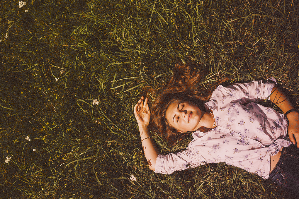 A woman is lying on the ground, surrounded by lush green grass, her eyes are closed, and she wears a gentle smile