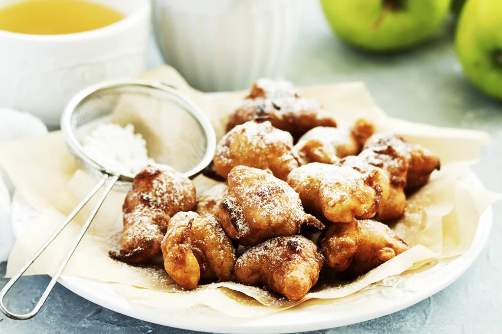 A plate overflowing with apple fritters sits on a table next to a powdered sugar shaker.