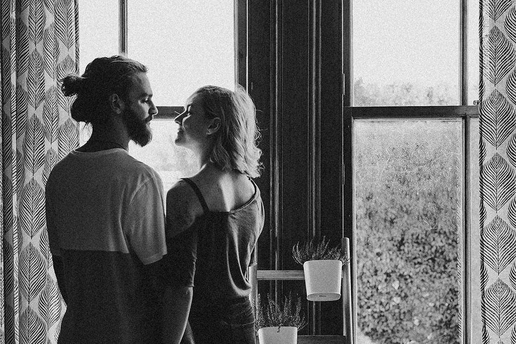 A man and a woman are holding each other's hands, looking at each other with smiles, while standing in front of a window in a room