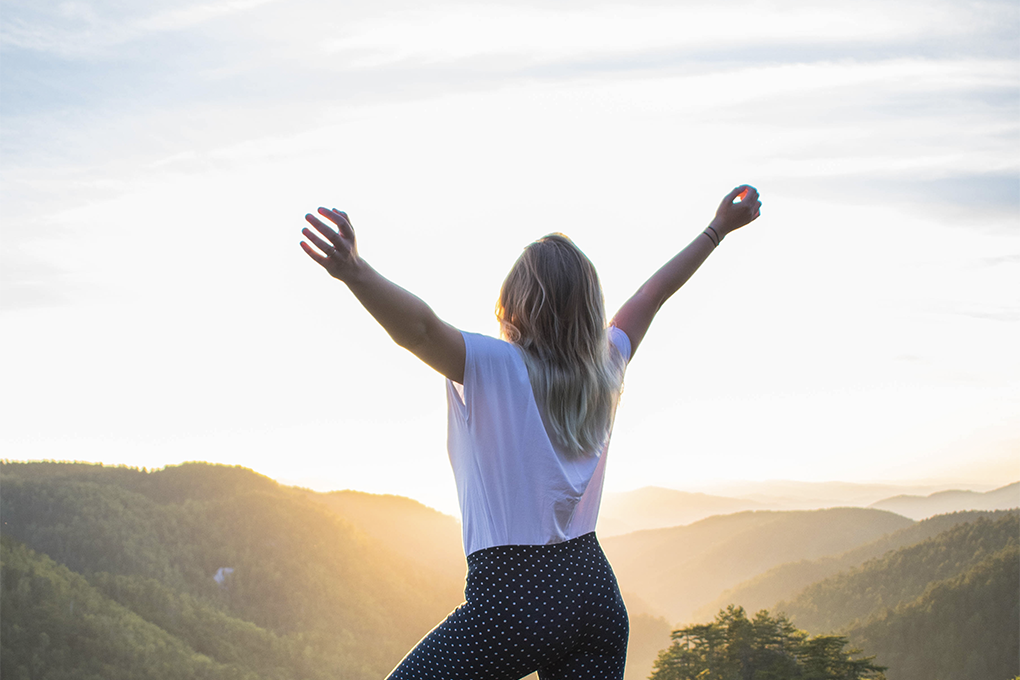 A woman, with he back facing the camera, holds her arms outstretched, as she looks into the distance at the sun over rolling hills.