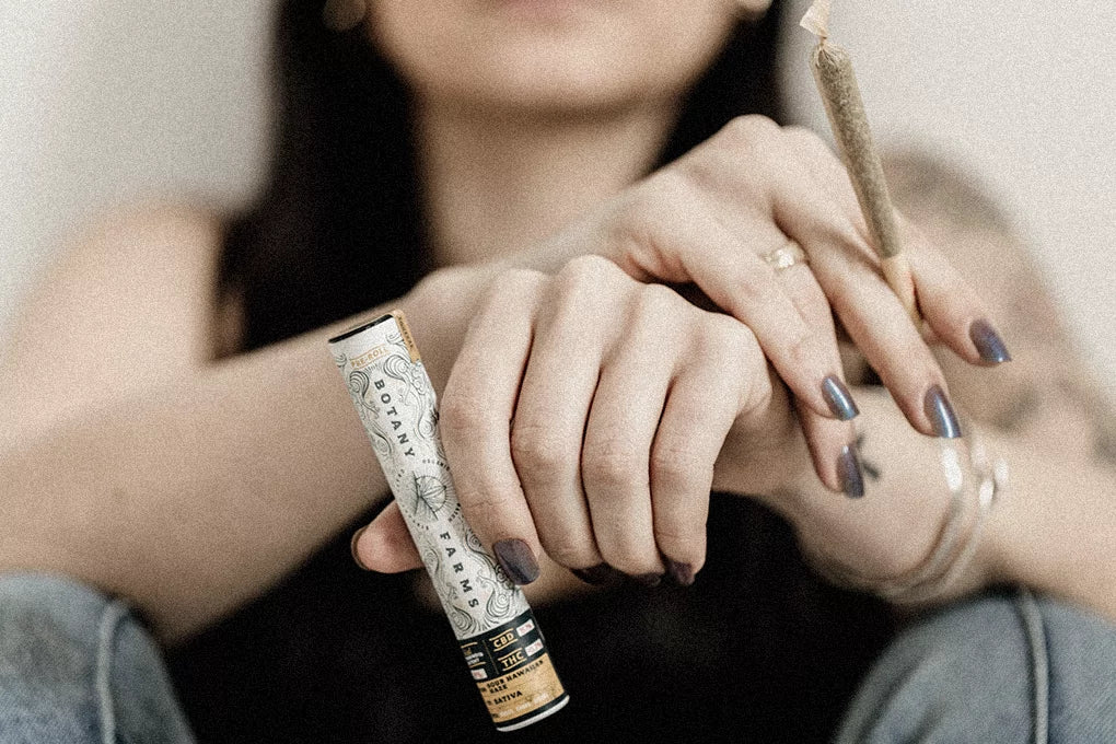 A person is seated on the floor with their hands crossed over their knees. In one hand they are holding a Delta 8 pre roll and in the other they are holding a Botany Farms preroll tube.