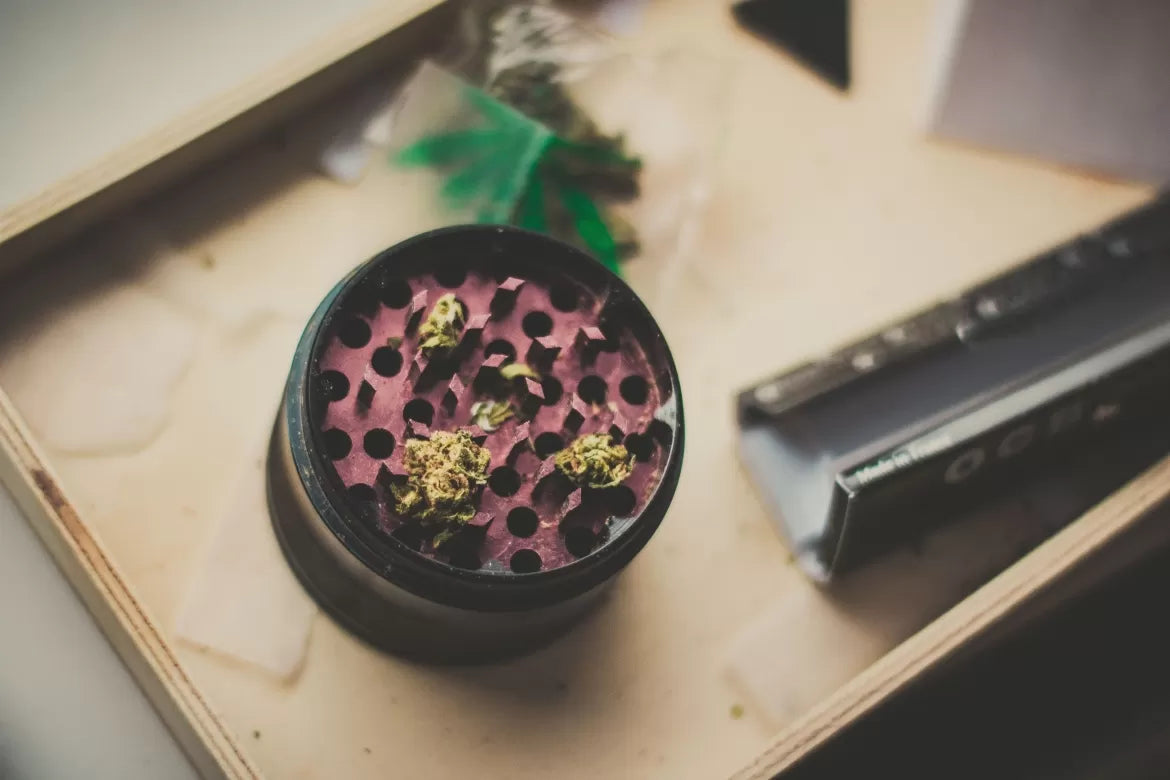 A cannabis grinder gilled with weed sits on a table beside rolling papers