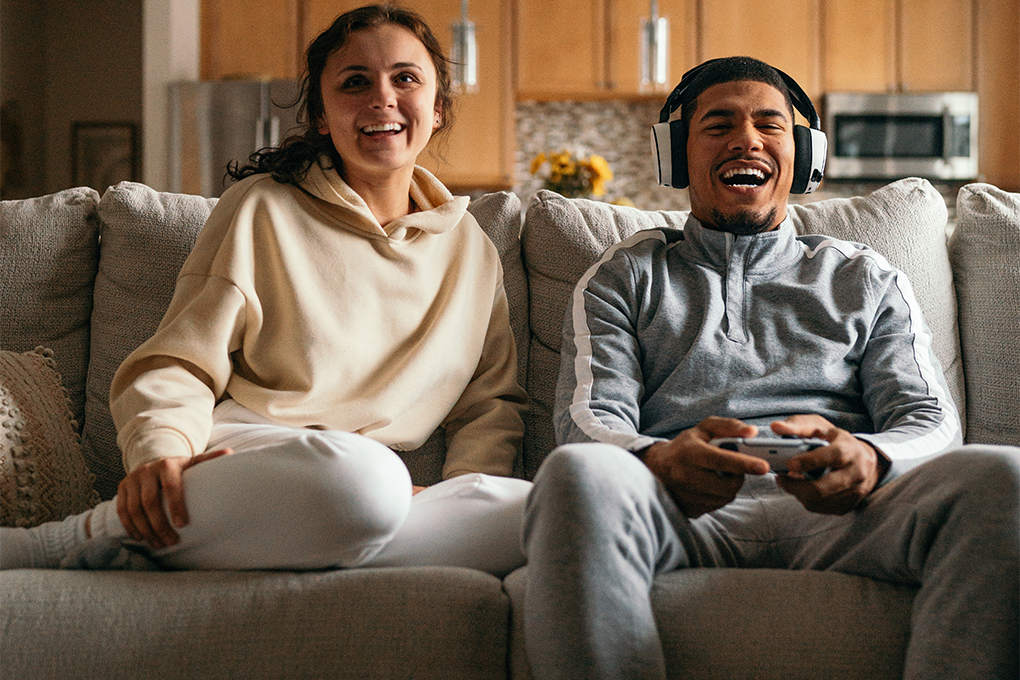A couple sitting on a sofa in their room, the boy wearing headphones and holding a gaming joystick, both laughing and enjoying the game