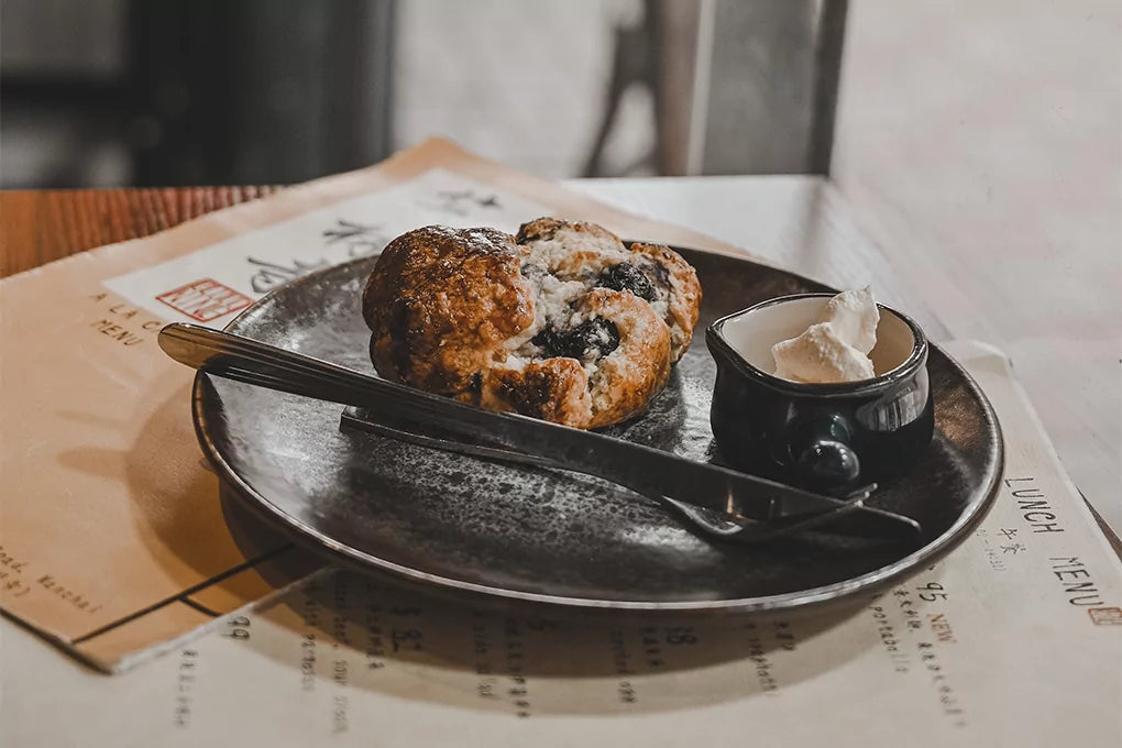 Freshly baked blueberry scone on white plate with silver fork and knife, placed beside papers on rustic wooden table