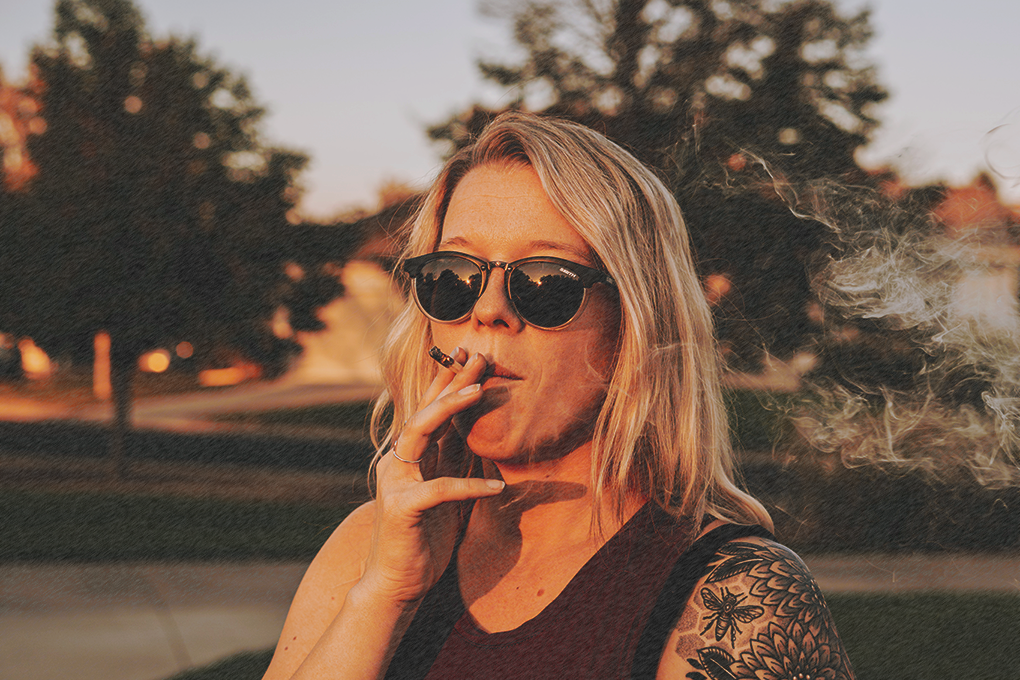 Woman with sunglasses enjoying a cannabis joint outdoors as sunlight illuminates her face