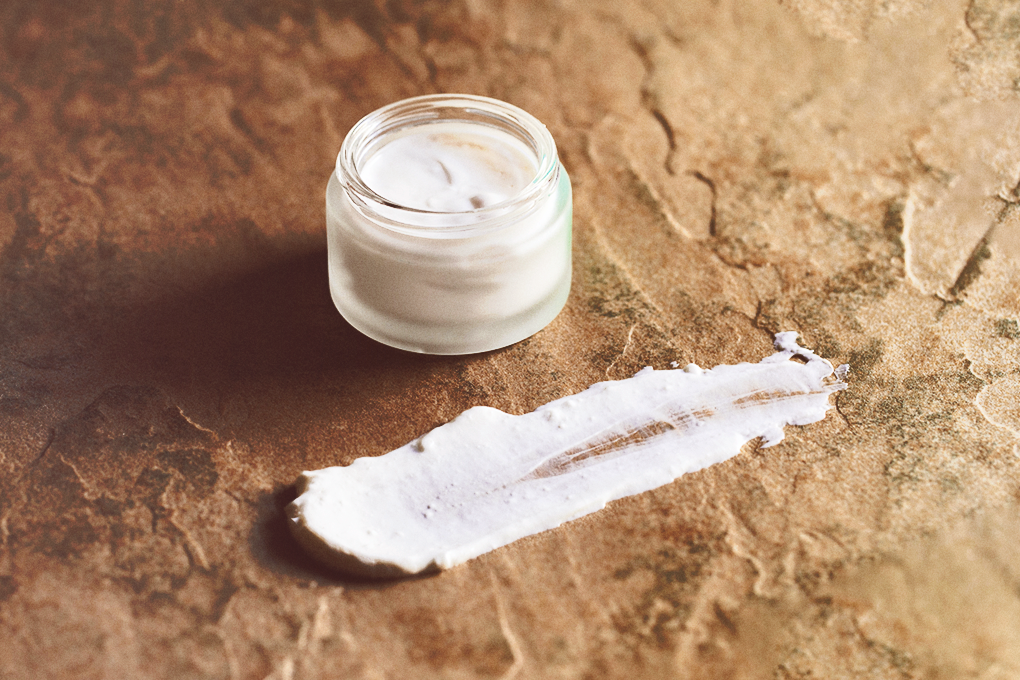A container of white CBD cream is positioned on a brown surface, with a portion of the cream smeared onto the surface