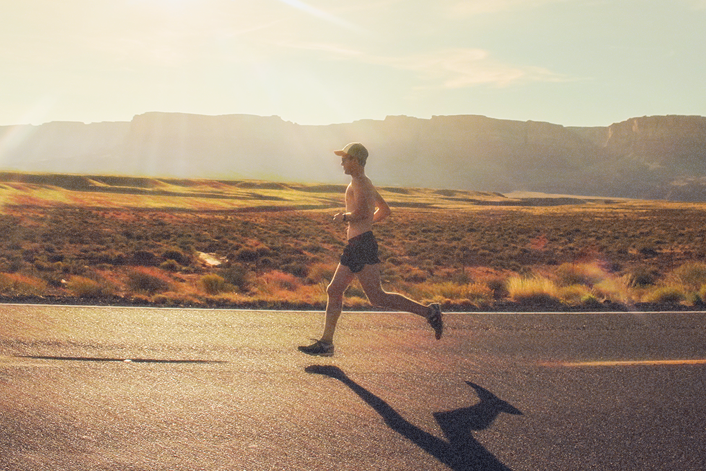 A man in sunglasses and hat jogging on the road with scenic mountains in the background on a sunny day