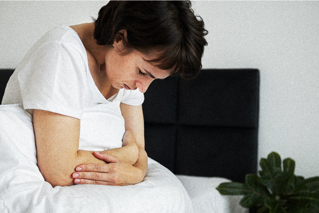 Woman on bed, hand on stomach, expressing discomfort, possibly experiencing stomach pain
