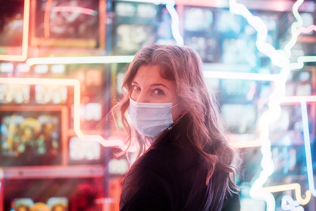 A woman with a covid 19 medical ask looks over her shoulder and into the camera. A colorful, lightning-like background stands behind her.