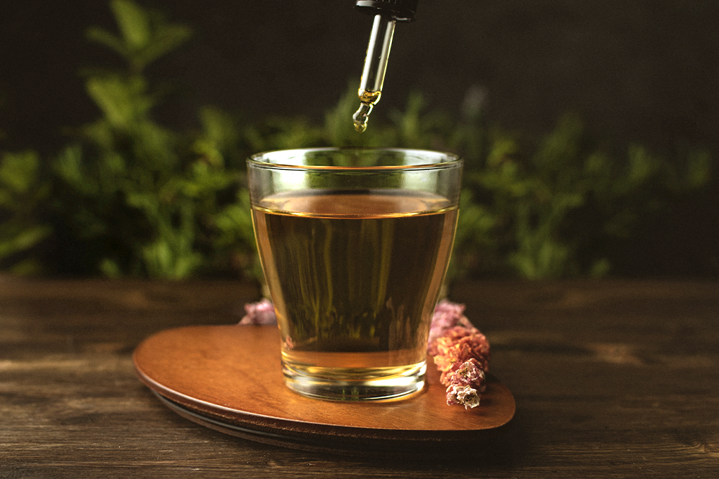 A clear glass of tea sits on a wooden coaster as a dropper of CBD hovers above it