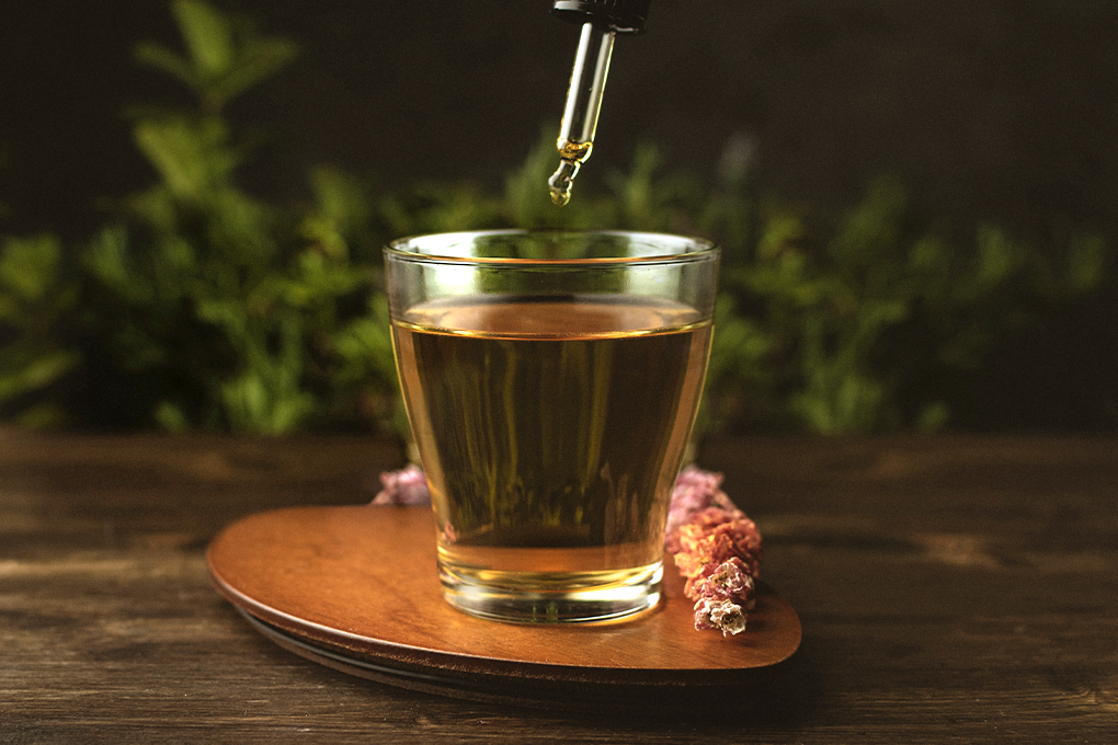 A glass jar containing CBD oil on a table, with a dropper releasing a drop of CBD oil into the jar.