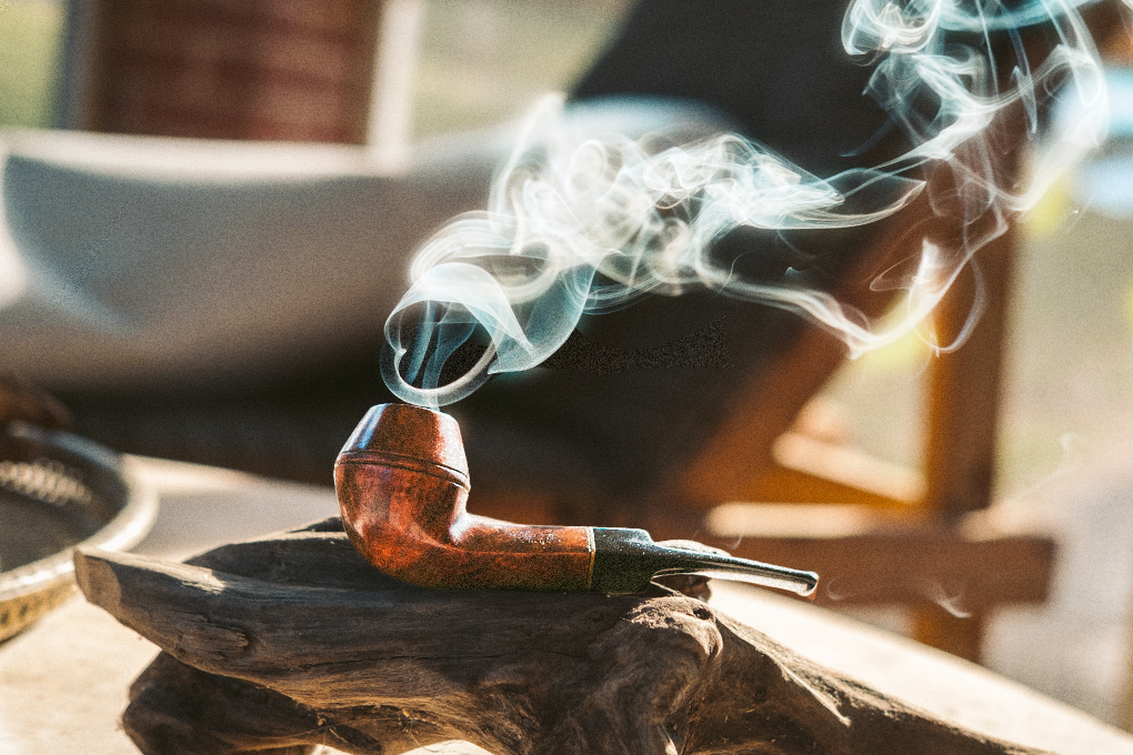 Smoke rises from briar wood tobacco pipe on wooden surface, with a wooden chair in the background against a hazy backdrop
