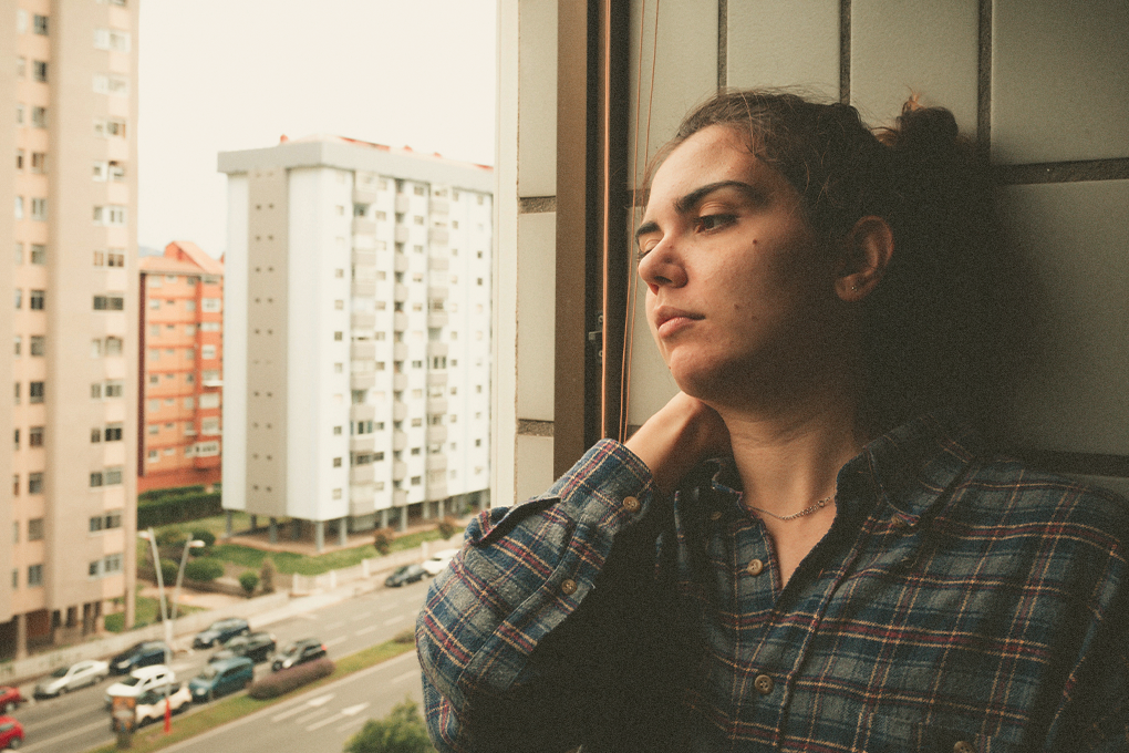 A tense-looking woman is sitting beside a window, looking out onto the street and There are some cars on the street and buildings on the other side
