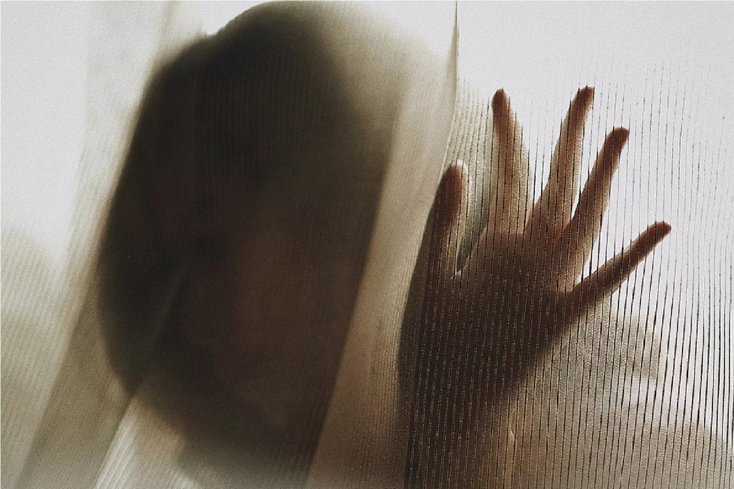 A woman in open hair standing in front of a sheer curtain with her face and hand touch the curtain