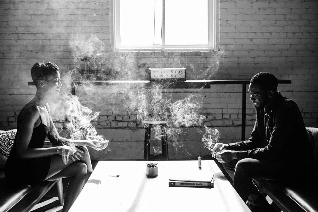 Two people sitting in a room together, with a table between them, enjoying CBD joints.