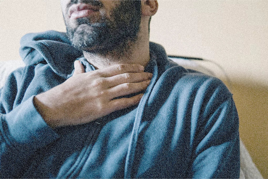 A closeup of a man in a blue sweater holding his throat with one hand to signify that he has a sore throat.