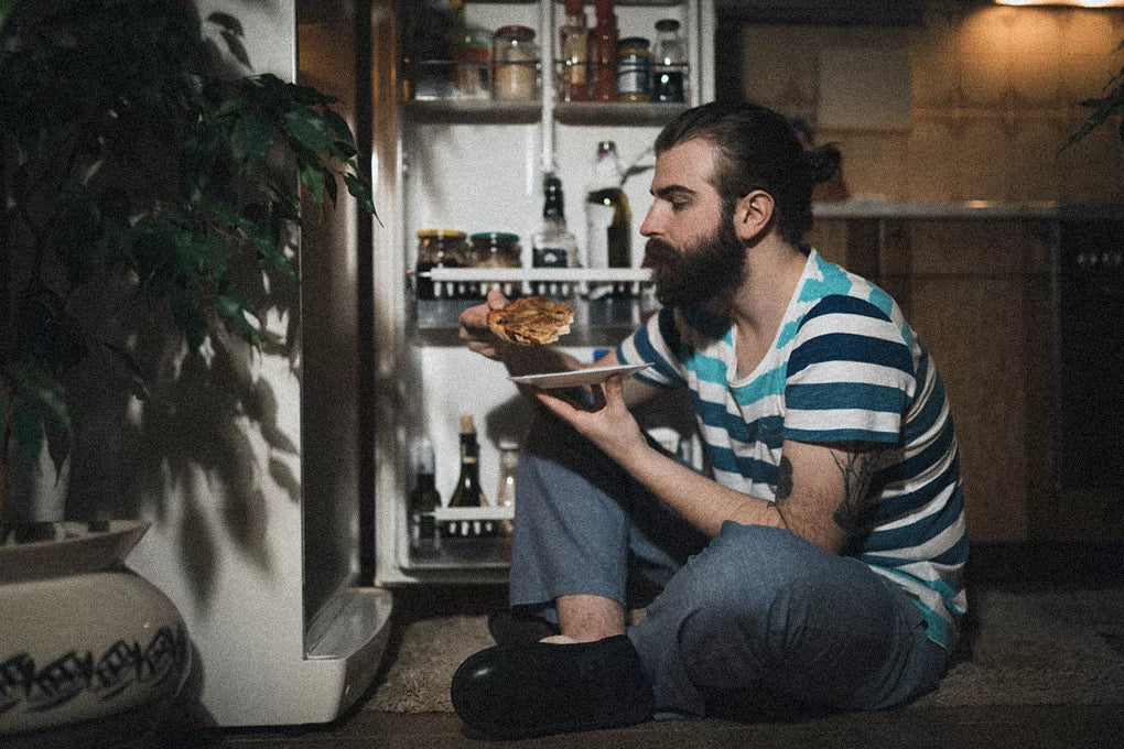 A person sits on the kitchen floor eating a pizza in front of an open fridge.