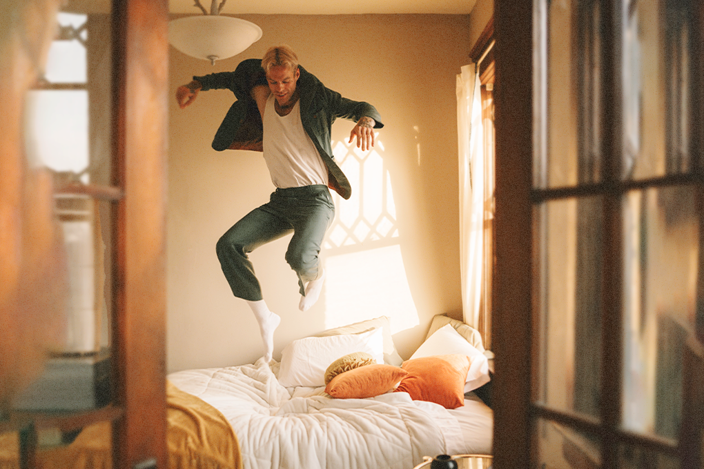 A boy in a shirt, jeans, and socks jumps energetically over the bed as if waking up and getting ready for the day