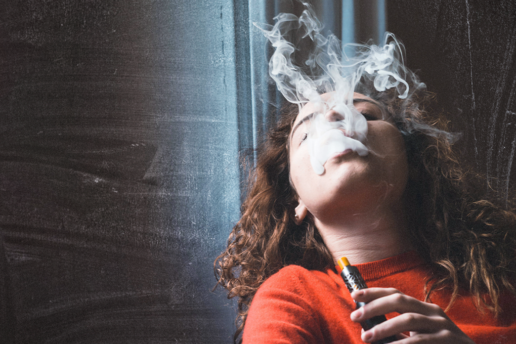 Woman exhaling vapor from a vape cartridge, eyes closed, head tilted back, against a backdrop of a black curtain