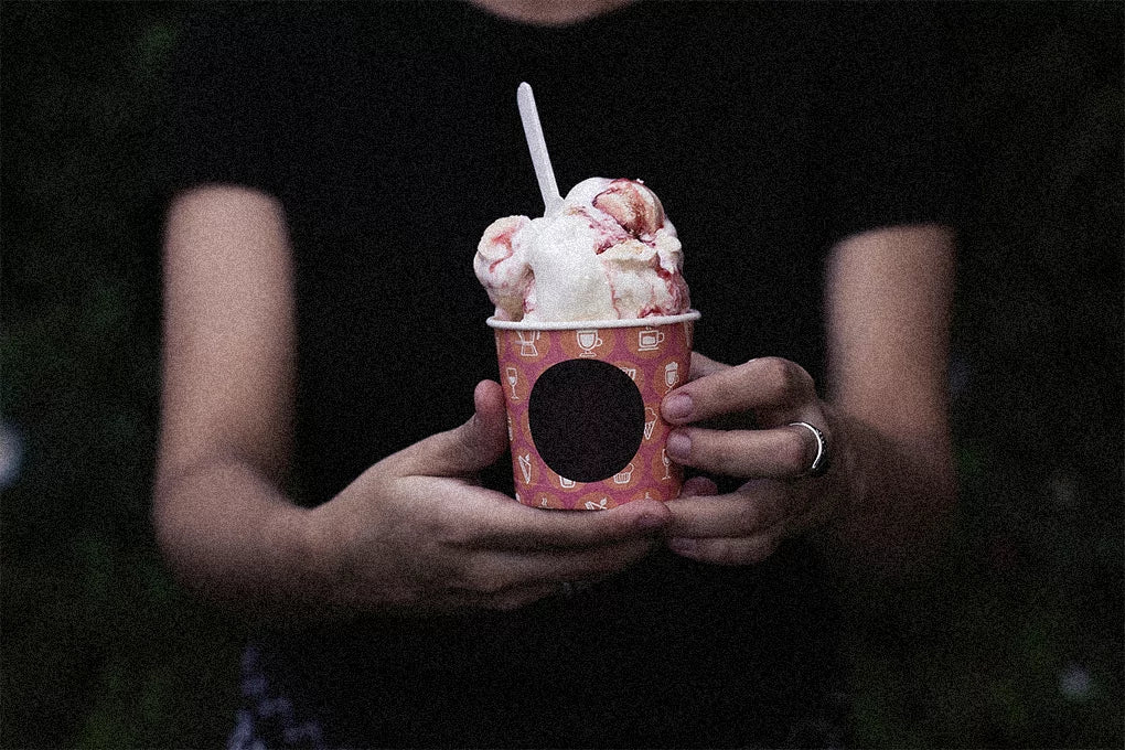 A pair of hands of a man wearing a black t-shirt, holding a paper cup of ice-cream with a white spoon