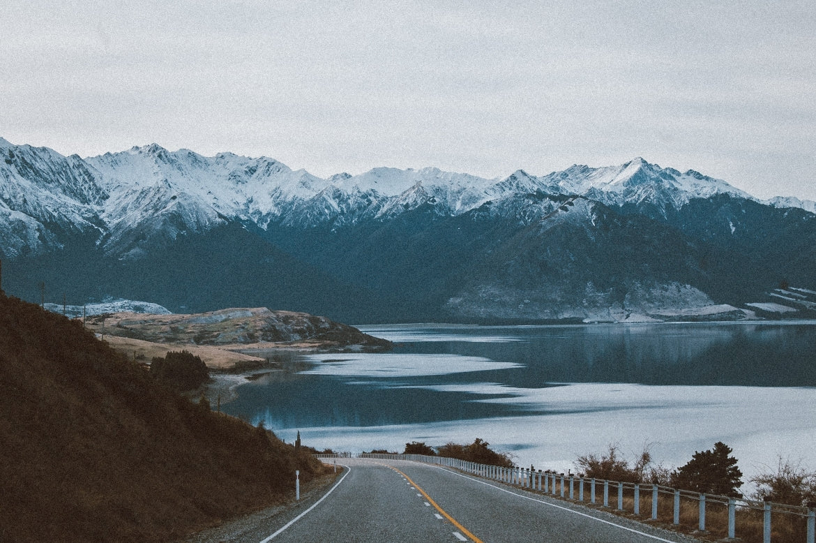 A road leads around a mountain side in Alaska. A large body of water and mountain peaks stand in the distance.
