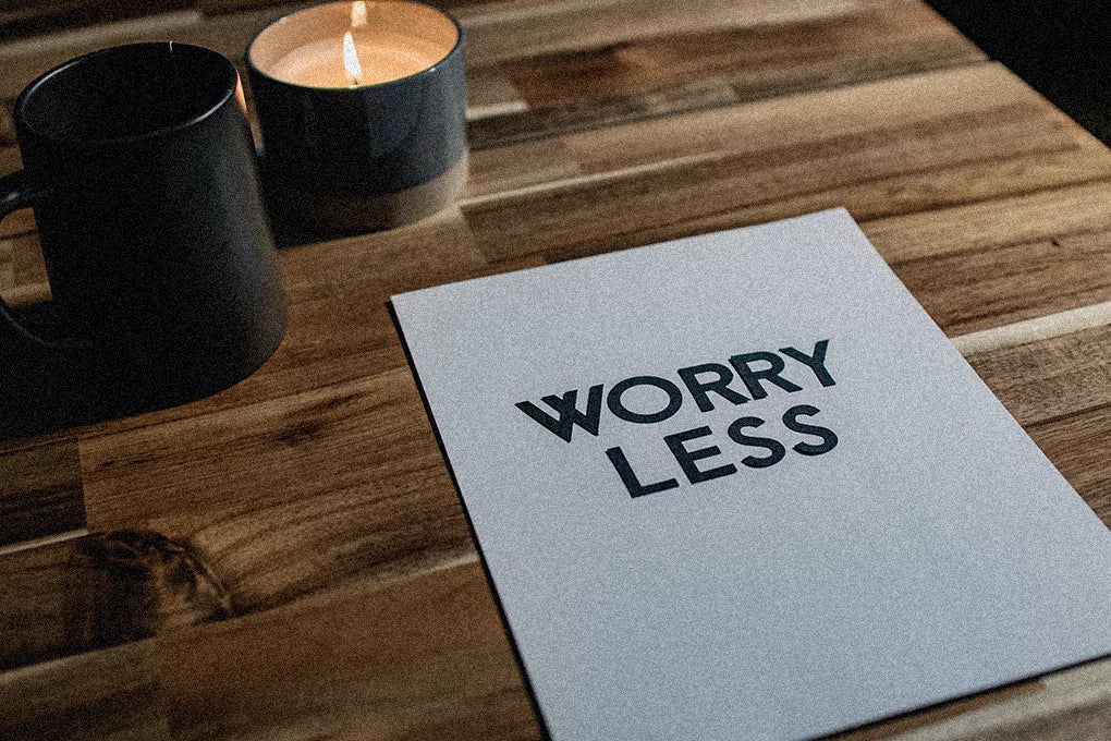 A piece of paper, with the words worry less written on it, sits on a wooden surface next to some lit herbal candles.