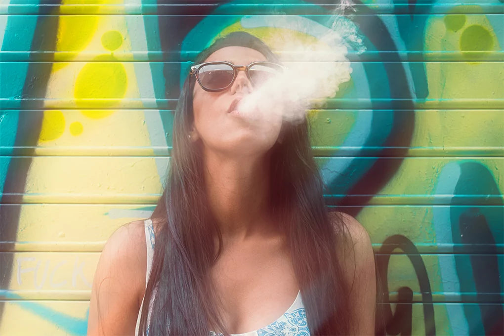 A woman wearing a sunglass looking up as she blows out smoke standing in front of a painted wall