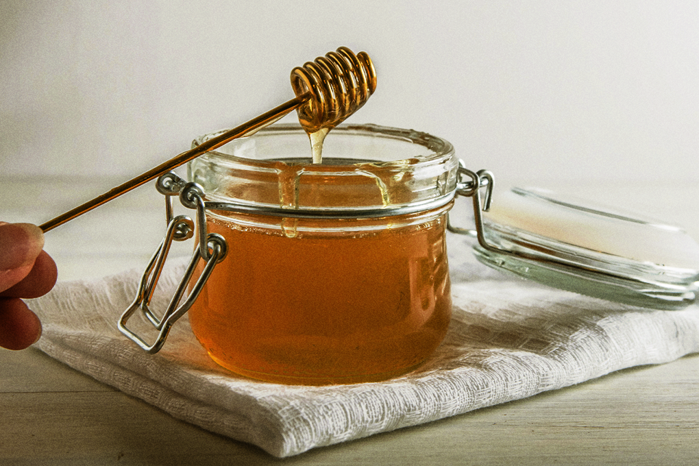 A close up shot of a honey jar with a honey dipper propped up against it