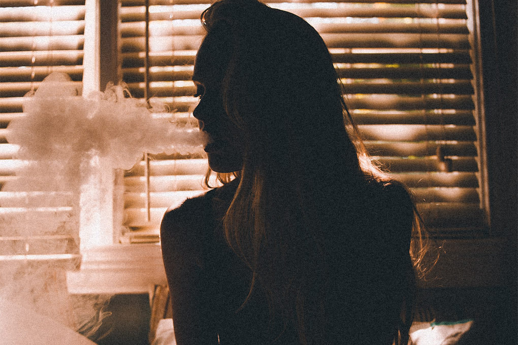 A woman in a dark room exhales smoke, with a closed window behind her, creating an atmospheric scene