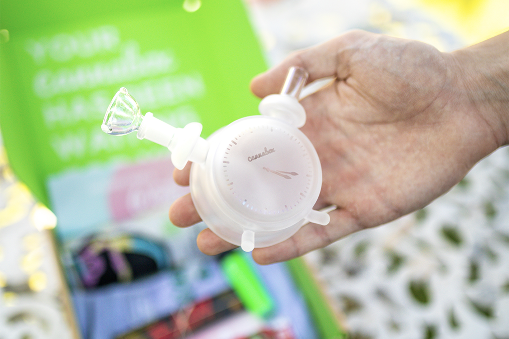 A close up shot of a hand holding a white silicone pipe shaped like a vintage alarm clock.