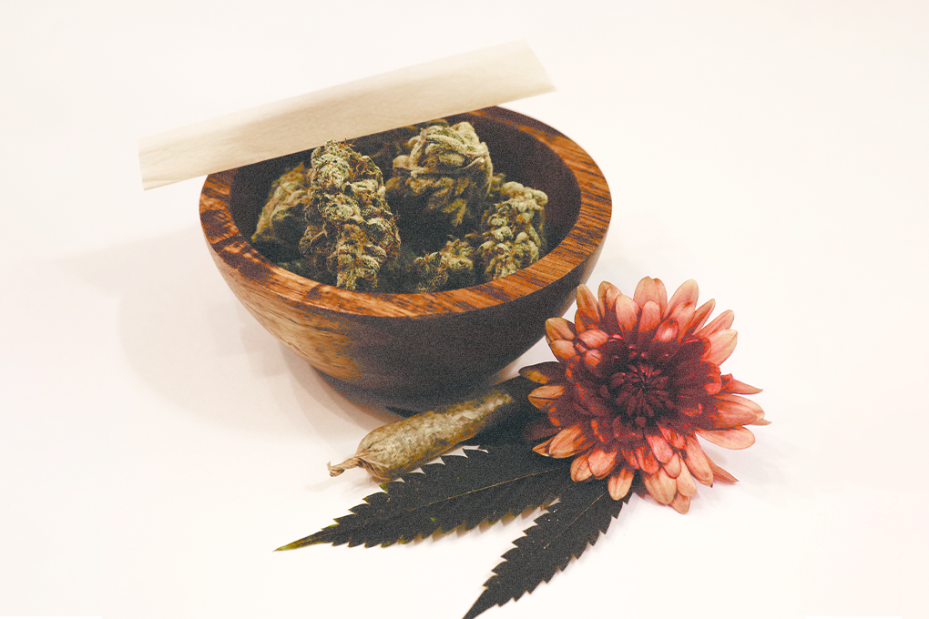 An arrangement featuring a small tub containing cannabis flowers, a rolled joint, a cannabis leaf, and a vibrant red flower placed beside the tub