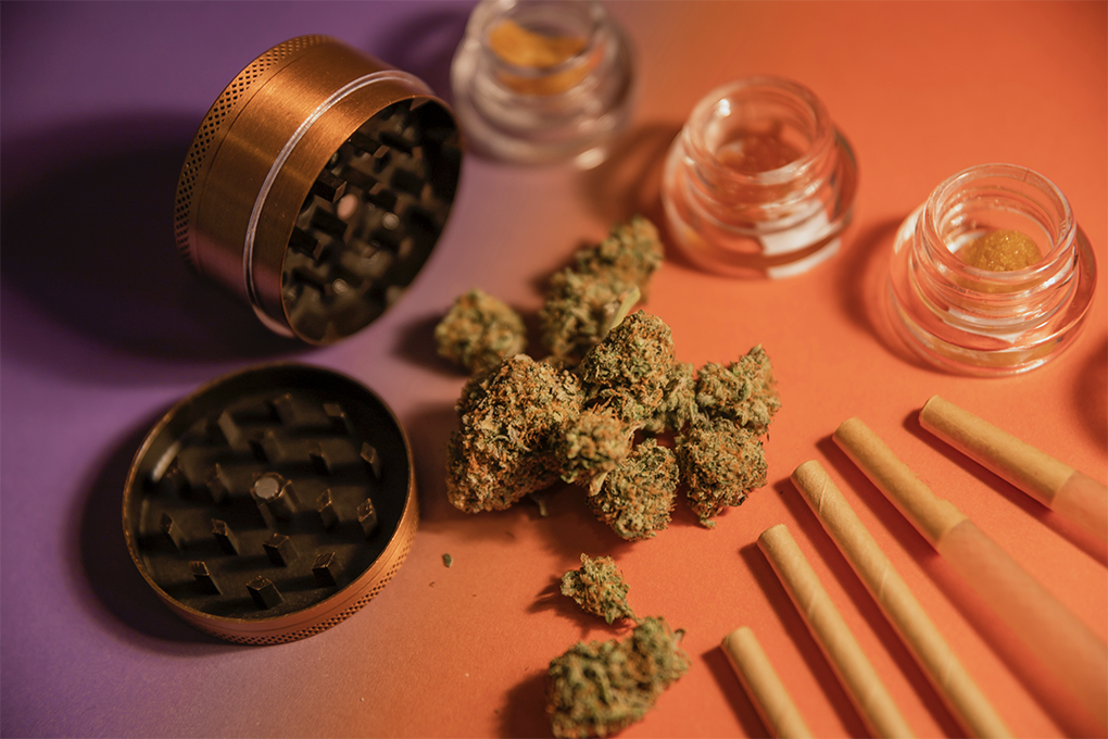 A weed grinder, some cannabis buds, pre rolls, and shatter jars are laid out on top of an orange surface.