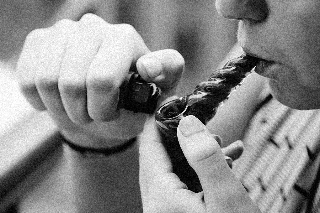 A black and white picture of a person lighting a bubbler pipe and preparing to smoke out of it.