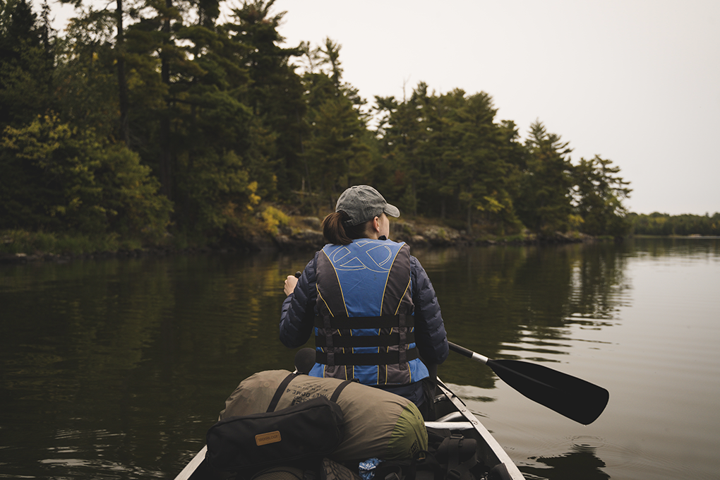 The view is from the back seat of a canoe. We can see the back of a man sitting in the front of the boat and paddling on the Mississippi river. in Minnesota