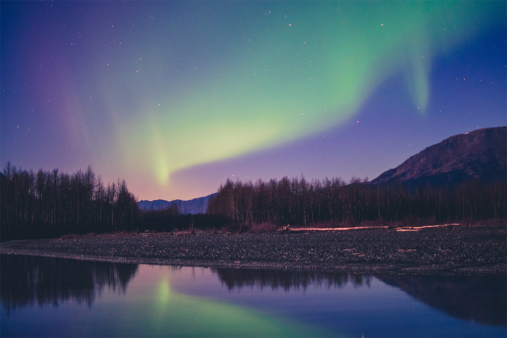 Alaska's evening sky glows with the Aurora Borealis, reflected in a quiet river with surrounding hills and trees
