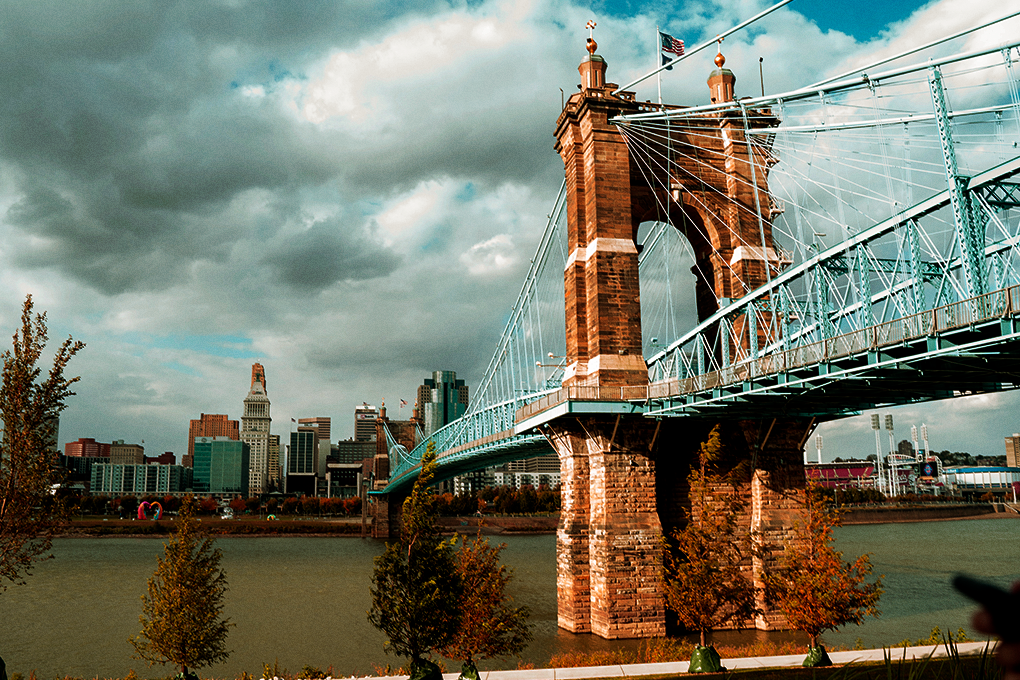 John A. Roebling Suspension Bridge spans the Ohio River, framed by a clear blue sky and drifting white clouds