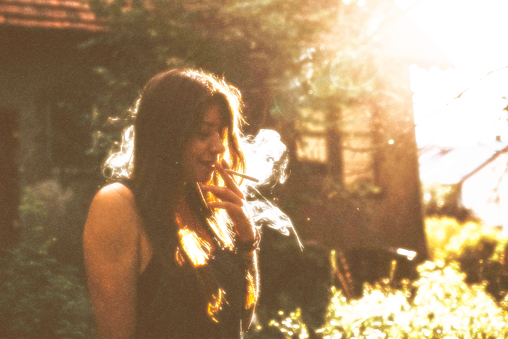 Smiling woman blissfully enjoying a joint, eyes closed, sunlight illuminating her hair, surrounded by trees and a house in the background