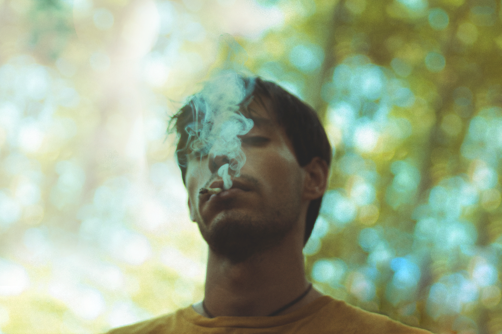 A man is seen outdoors, exhaling smoke from a joint, while his closed eyes indicate a state of intoxication