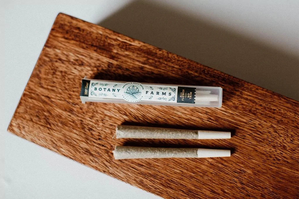 Two Botany Farms HHC pre rolls sit on a wooden surface next to a Botany Farms HHC pre roll tube.