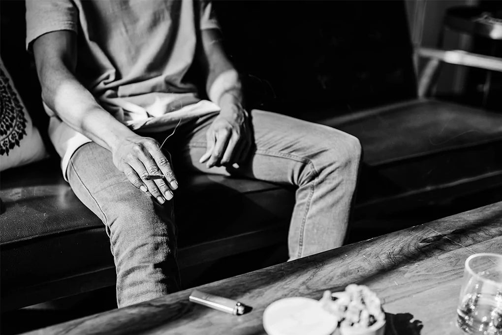 A black and white image of a man sitting on a couch holding an infused pre roll joint between his fingers.