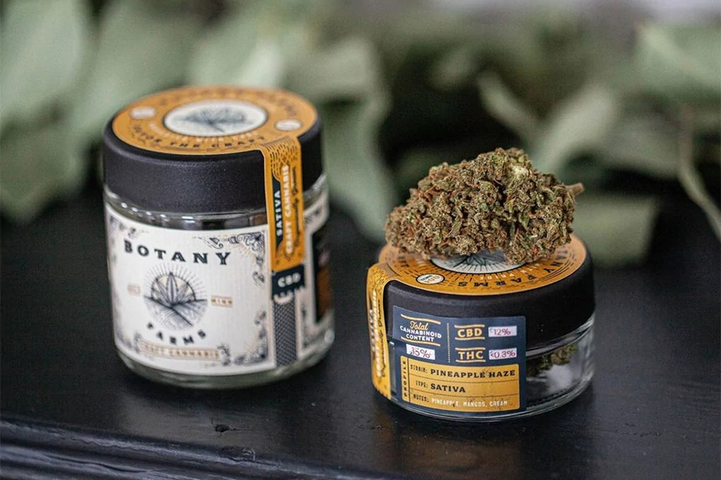 Two jars of Botany Farms flower sit atop a black surface. One of the jars has a cannabis bud on top of it.
