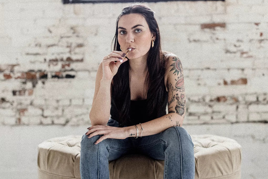 Young woman sitting on a sofa in front of a white wall, holding a cartridge in her right hand up to her mouth with a tattoo on her left hand