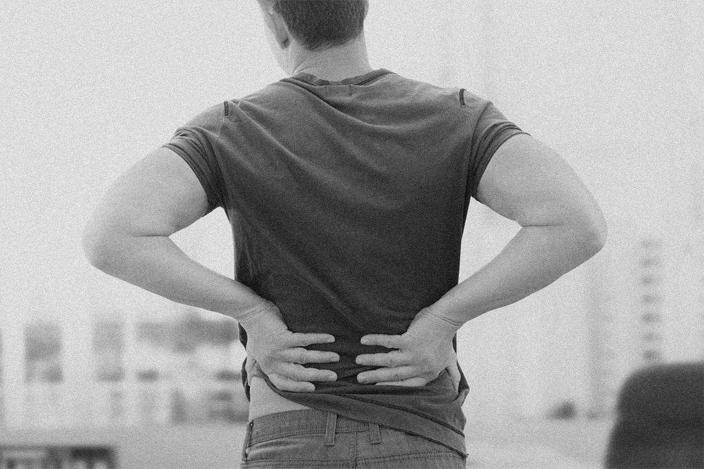 A black and white image of a man facing away from the camera as he holds his lower back with both hands signifying that he is in pain.