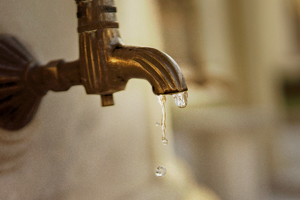 A water drop falls from a golden-colored water tap against a hazy background