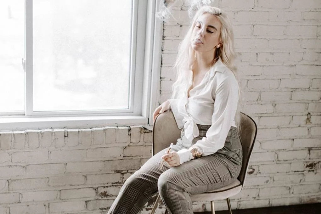 A woman sits on a chair next to a window and smokes from a Delta-8 TCH cartridge.