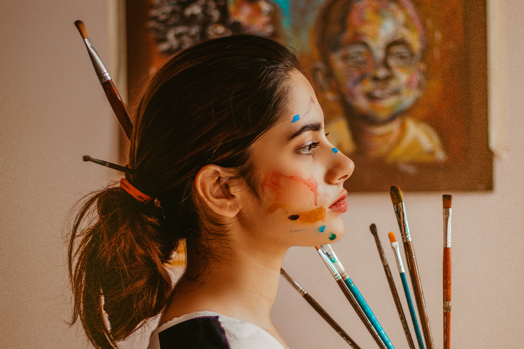 A side profile of an artist holding up brushes. Her face is covered in paint.
