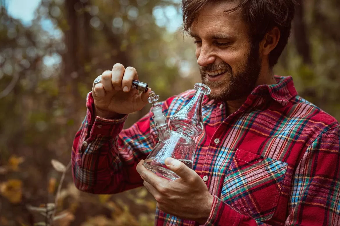 A man in a plaid shirt stands in the forest and enjoys hemp flower from a bong to help his anxiety.